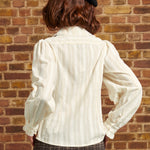 Back view of an ivory Victorian-inspired blouse with textured vertical stripes, long puff sleeves, and ruffled cuffs, styled with plaid trousers and a black beret.