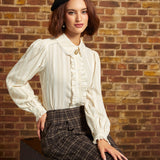 A woman wearing a cream pleated long-sleeve blouse with ruffles, a black beret, and brown plaid trousers, posing on a vintage trunk against a brick wall.