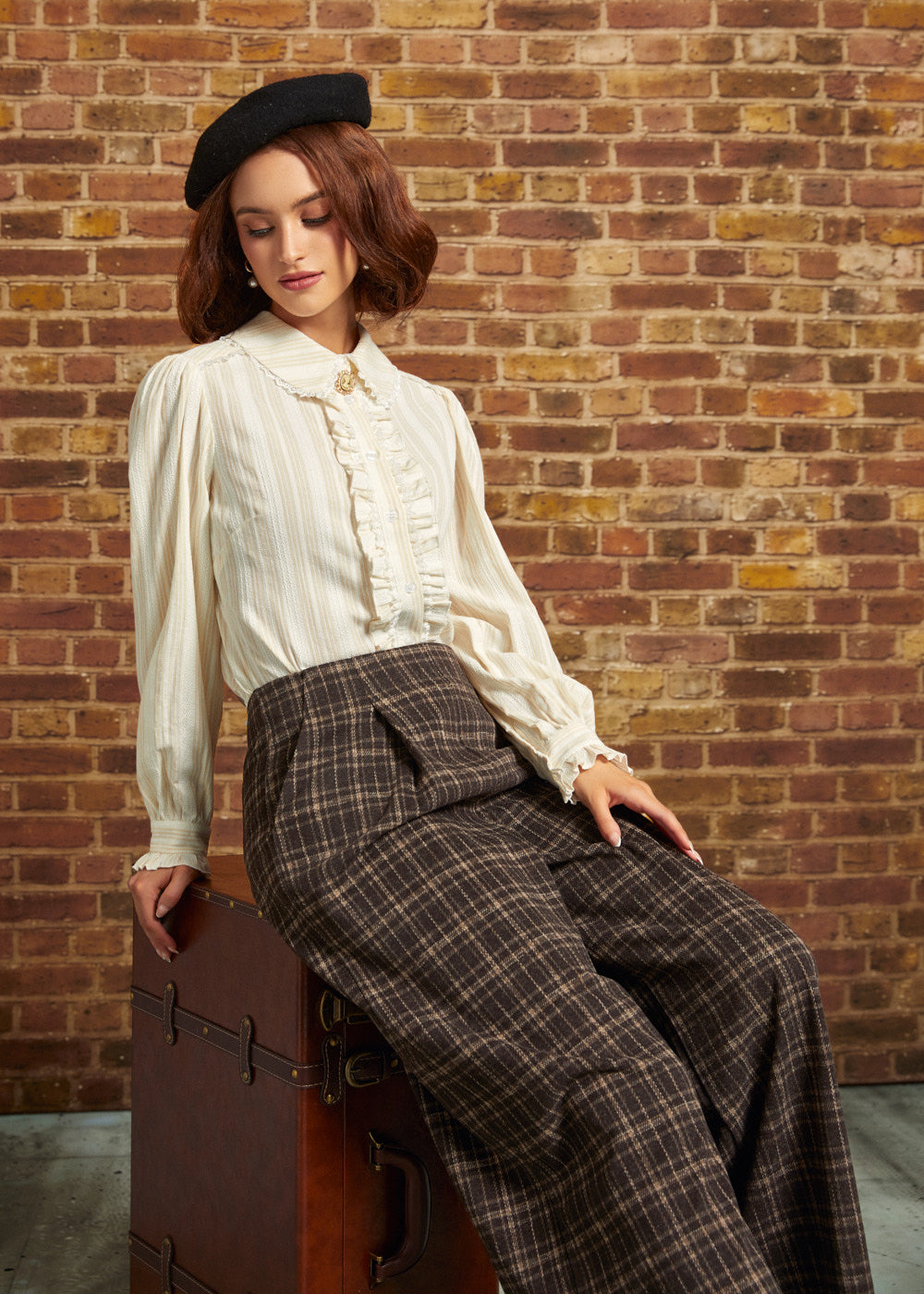 A woman wearing a cream ruffled blouse, brown plaid wide-leg trousers, and a black beret sits on a vintage brown trunk against a brick wall.