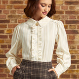 A woman wearing a cream ruffled long-sleeve blouse with a high collar, paired with dark brown plaid trousers and a black beret, standing against a brick wall.