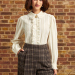 A woman wearing a cream ruffled blouse, brown plaid trousers, and a black beret stands in front of a brick wall.