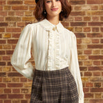 A woman wearing a cream pleated long-sleeve blouse with a ruffled collar and a gold cameo accent, paired with brown plaid trousers and a black beret, standing against a brick wall.
