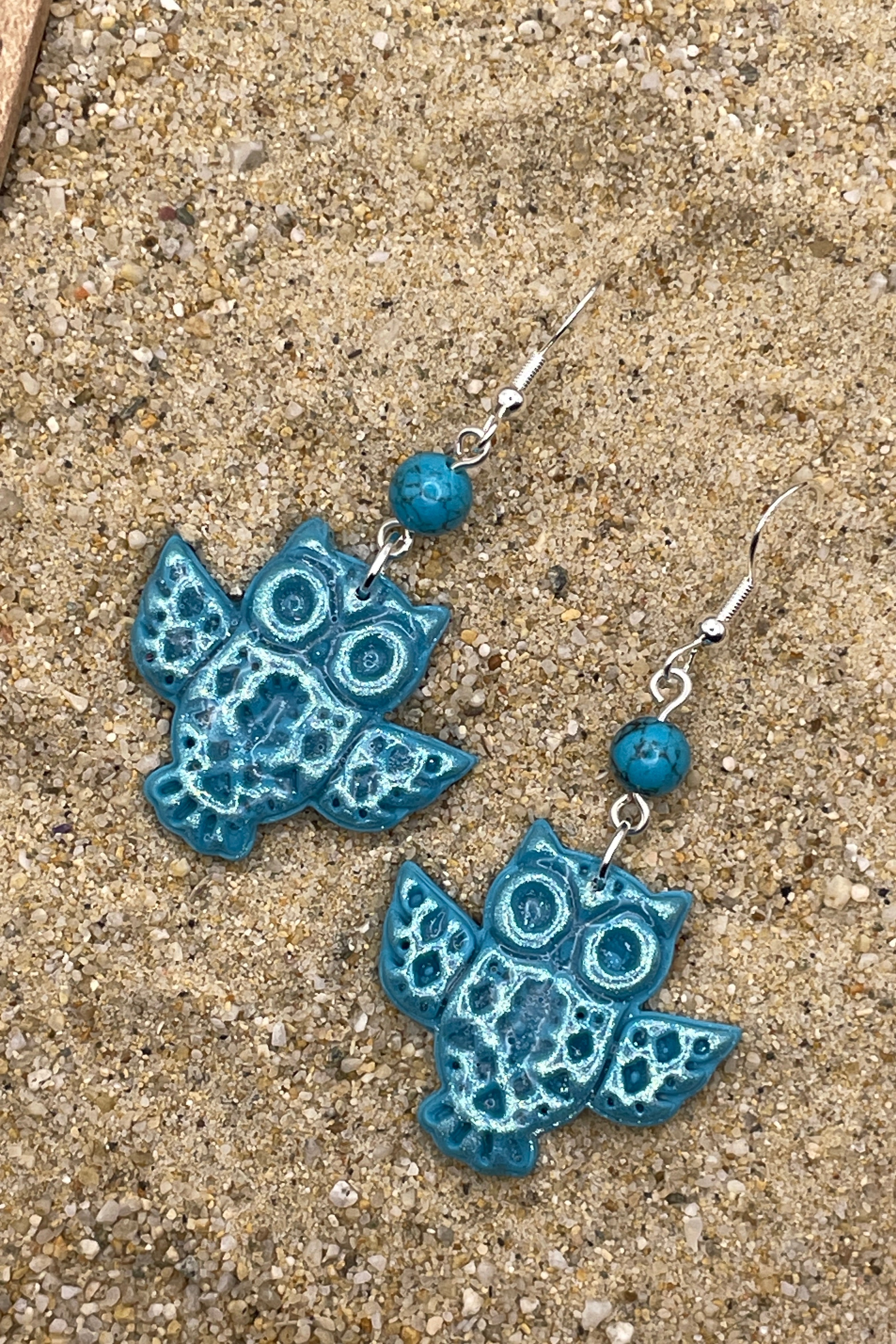 Blue owl-shaped earrings on a sandy surface