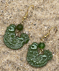 Green chicken shaped earrings with gold hooks on a sandy surface