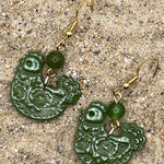 Green chicken shaped earrings with gold hooks on a sandy surface