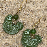 Green chicken shaped earrings with gold hooks on a sandy surface