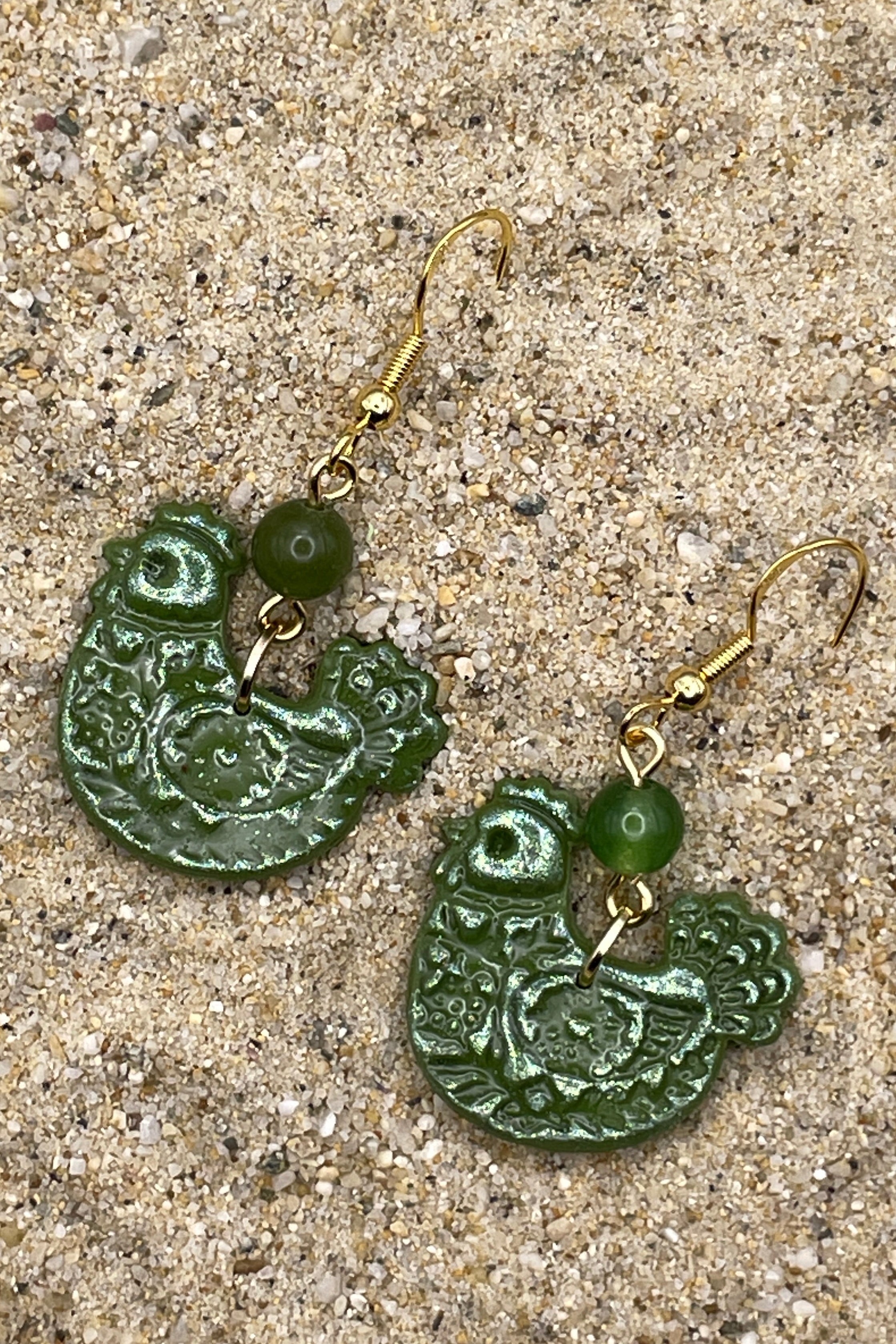 Green chicken shaped earrings with gold hooks on a sandy surface