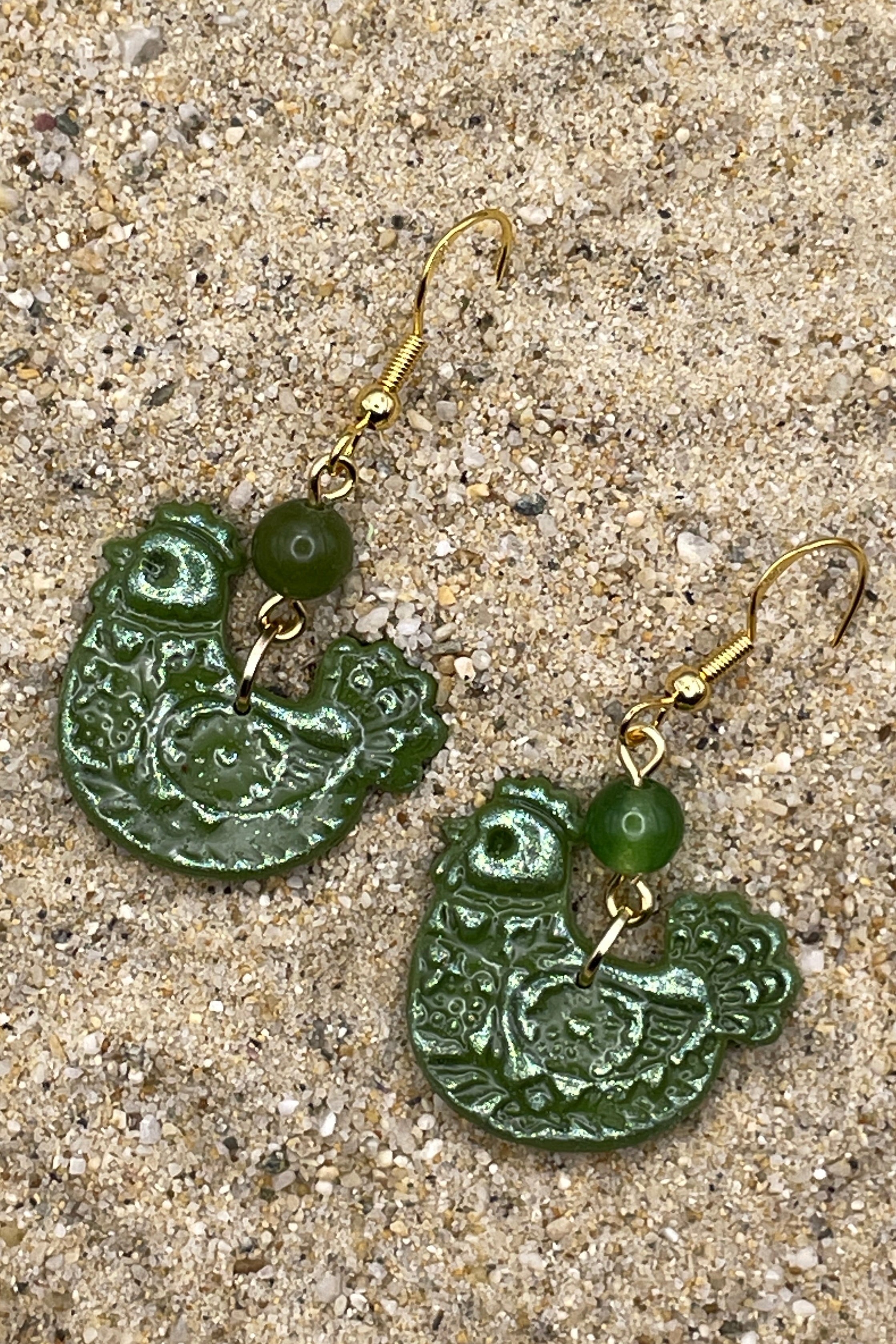 Green chicken shaped earrings with gold hooks on a sandy surface