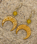 Gold crescent moon earrings with yellow beads on a sandy surface