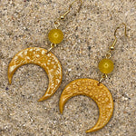 Gold crescent moon earrings with yellow beads on a sandy surface