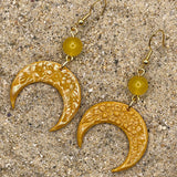 Gold crescent moon earrings with yellow beads on a sandy surface