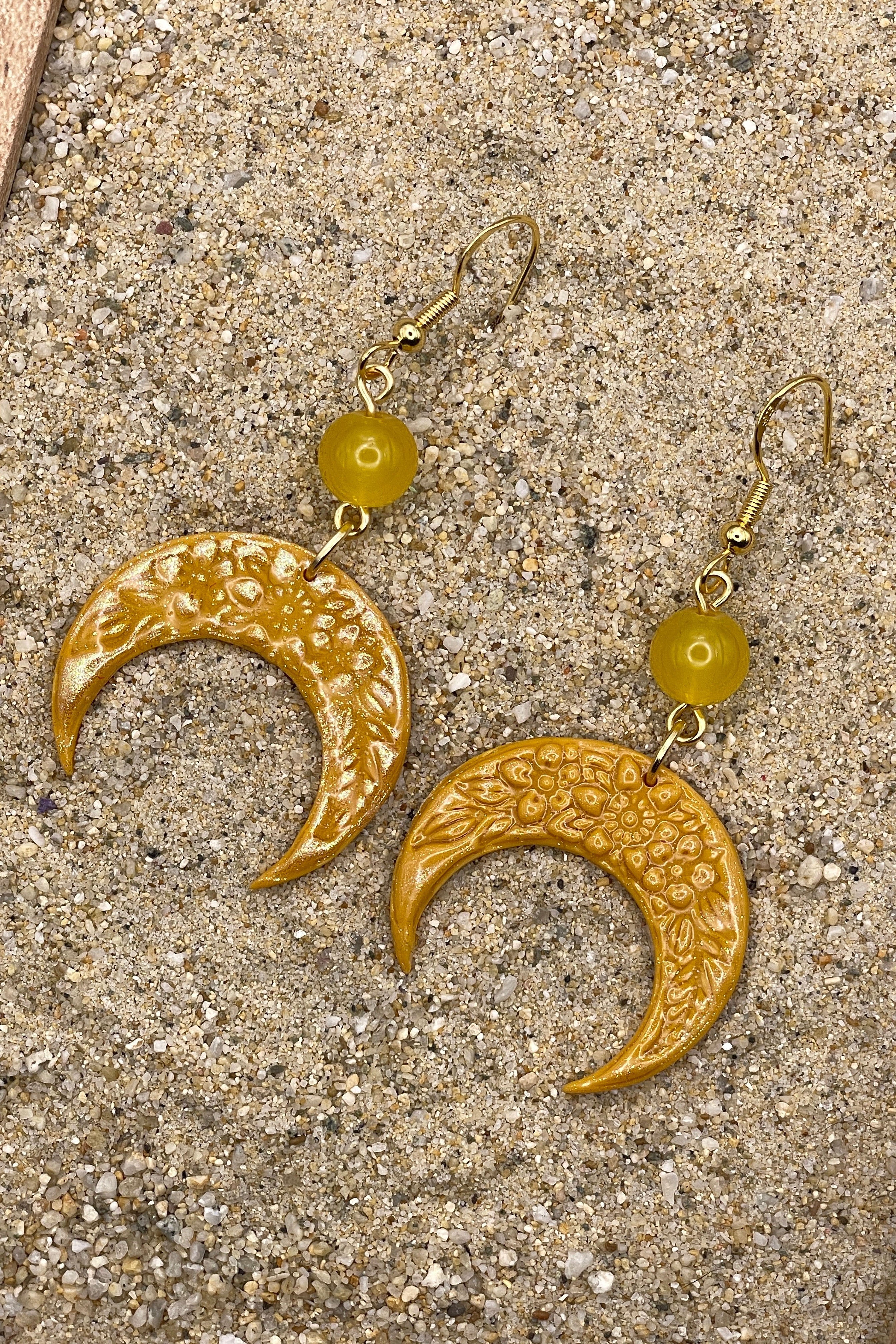 Gold crescent moon earrings with yellow beads on a sandy surface