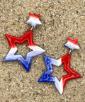 Star-shaped earrings in red, white, and blue on a sandy surface