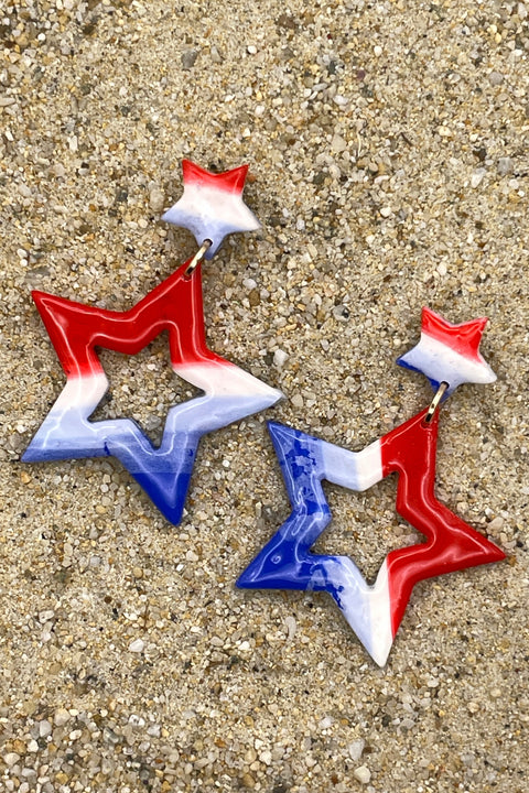 Star-shaped earrings in red, white, and blue on a sandy surface