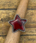 A star-shaped silver ring with a deep red cabochon stone, displayed on a wooden dowel over a rustic wooden plank background.