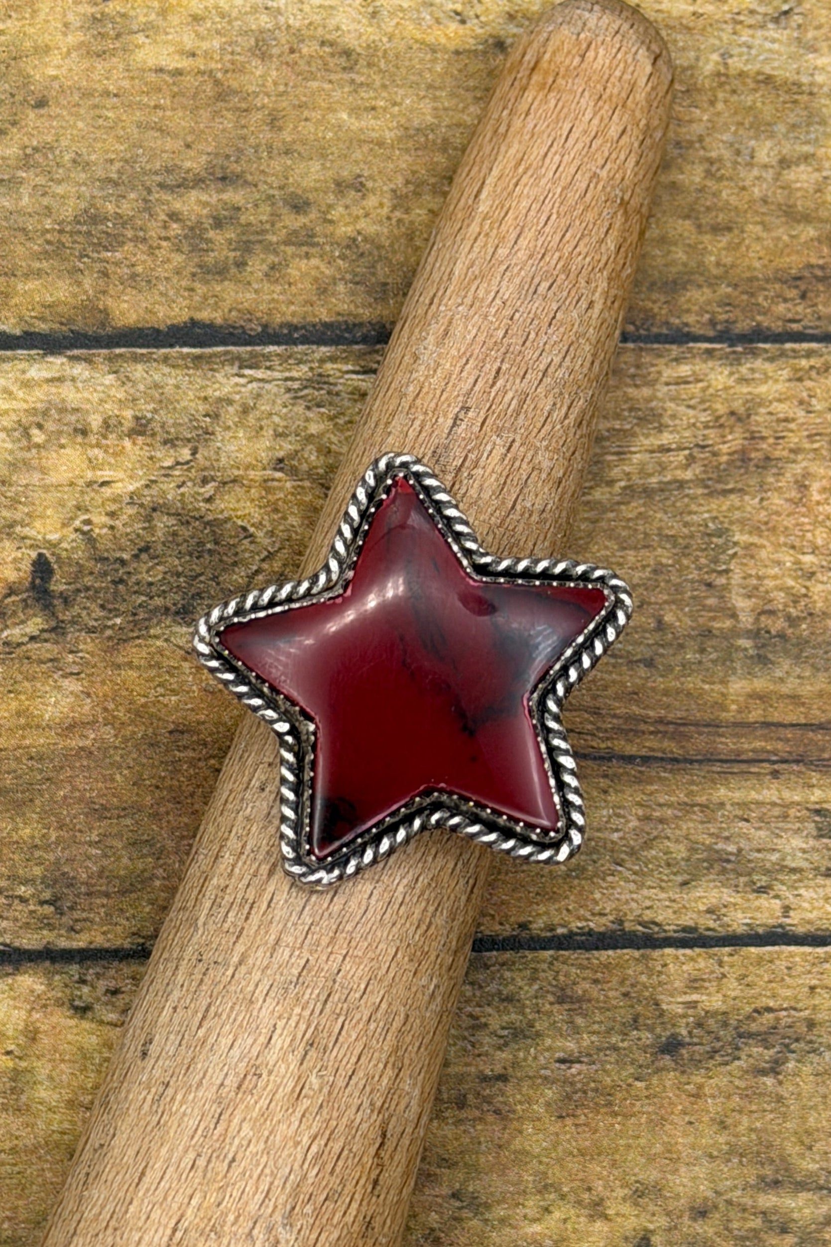 A star-shaped silver ring with a deep red cabochon stone, displayed on a wooden dowel over a rustic wooden plank background.