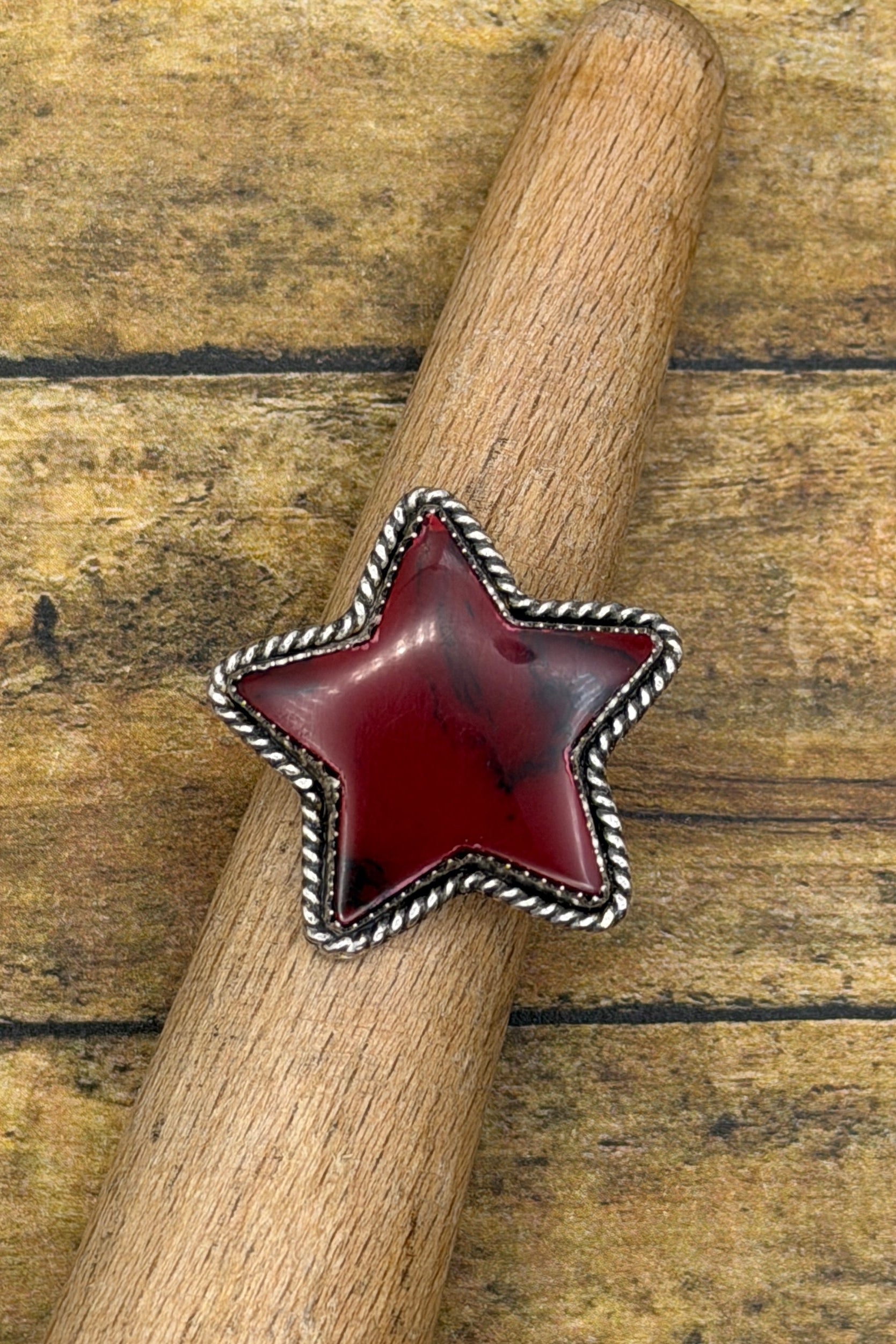 A star-shaped silver ring with a deep red cabochon stone, displayed on a wooden dowel over a rustic wooden plank background.