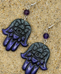 A pair of Hamsa hand dangle earrings with a glossy black and deep purple gradient hand design, a small purple bead, and silver fishhook wires, displayed on a sandy surface.
