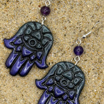 A pair of Hamsa hand dangle earrings with a glossy black and deep purple gradient hand design, a small purple bead, and silver fishhook wires, displayed on a sandy surface.