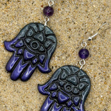 A pair of Hamsa hand dangle earrings with a glossy black and deep purple gradient hand design, a small purple bead, and silver fishhook wires, displayed on a sandy surface.