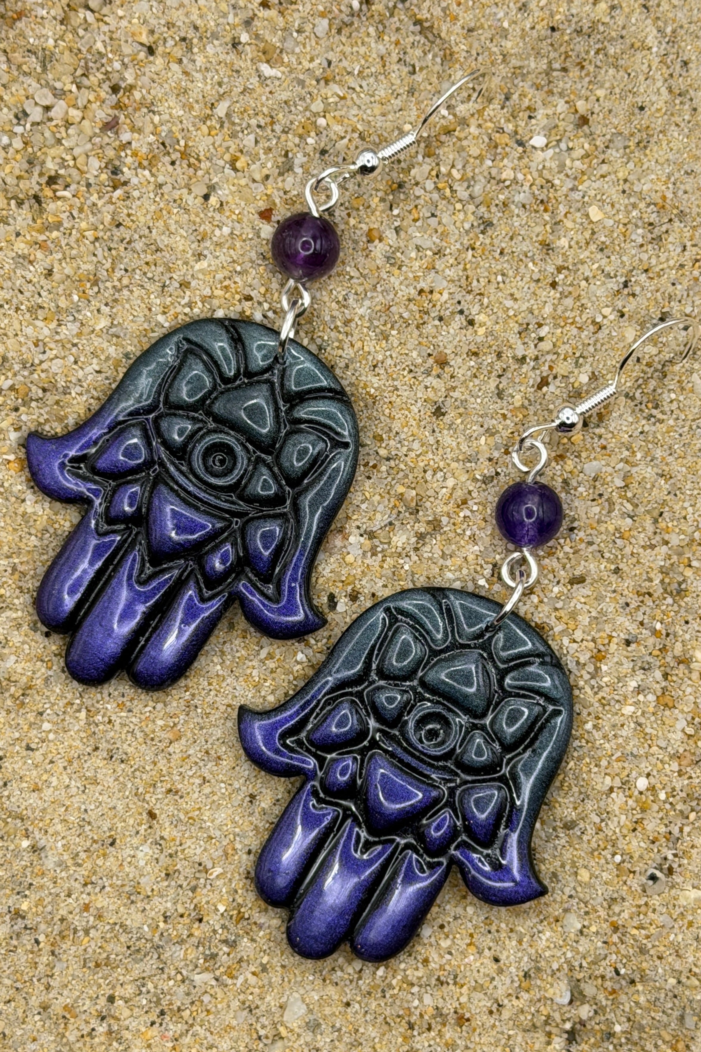 A pair of Hamsa hand dangle earrings with a glossy black and deep purple gradient hand design, a small purple bead, and silver fishhook wires, displayed on a sandy surface.