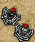 A pair of gothic spiderweb dangle earrings with black and white web patterns, red roses, green leaves, and a central black oval containing 'RIP' text, displayed on a sandy surface.