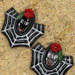 A pair of gothic spiderweb dangle earrings with black and white web patterns, red roses, green leaves, and a central black oval containing 'RIP' text, displayed on a sandy surface.