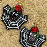 A pair of gothic spiderweb dangle earrings with black and white web patterns, red roses, green leaves, and a central black oval containing 'RIP' text, displayed on a sandy surface.