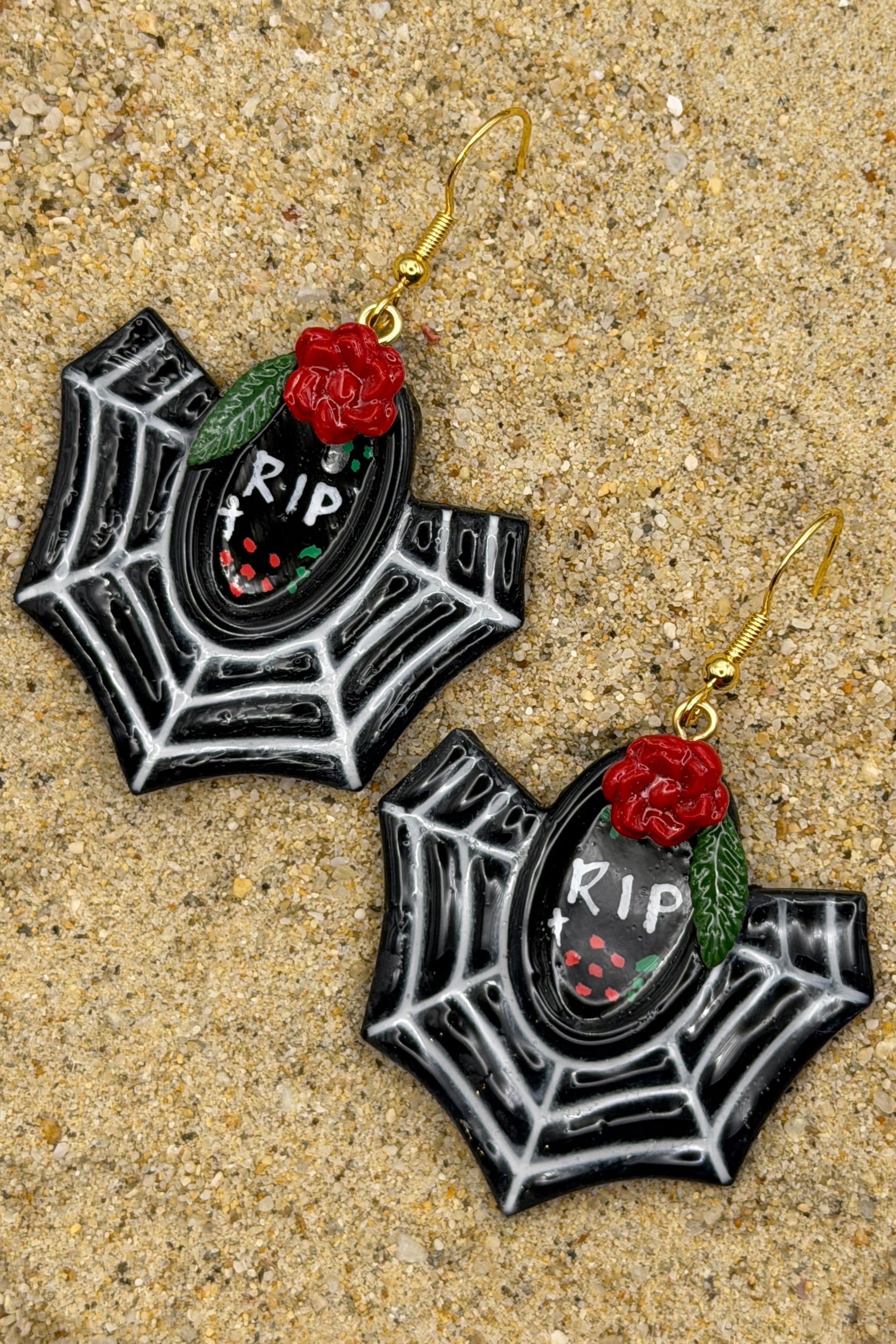 A pair of gothic spiderweb dangle earrings with black and white web patterns, red roses, green leaves, and a central black oval containing 'RIP' text, displayed on a sandy surface.