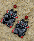 A pair of black vampire-shaped dangle earrings with red accents, silver eyes, and gold fishhook wires, displayed on a sandy surface.