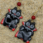 A pair of black vampire-shaped dangle earrings with red accents, silver eyes, and gold fishhook wires, displayed on a sandy surface.