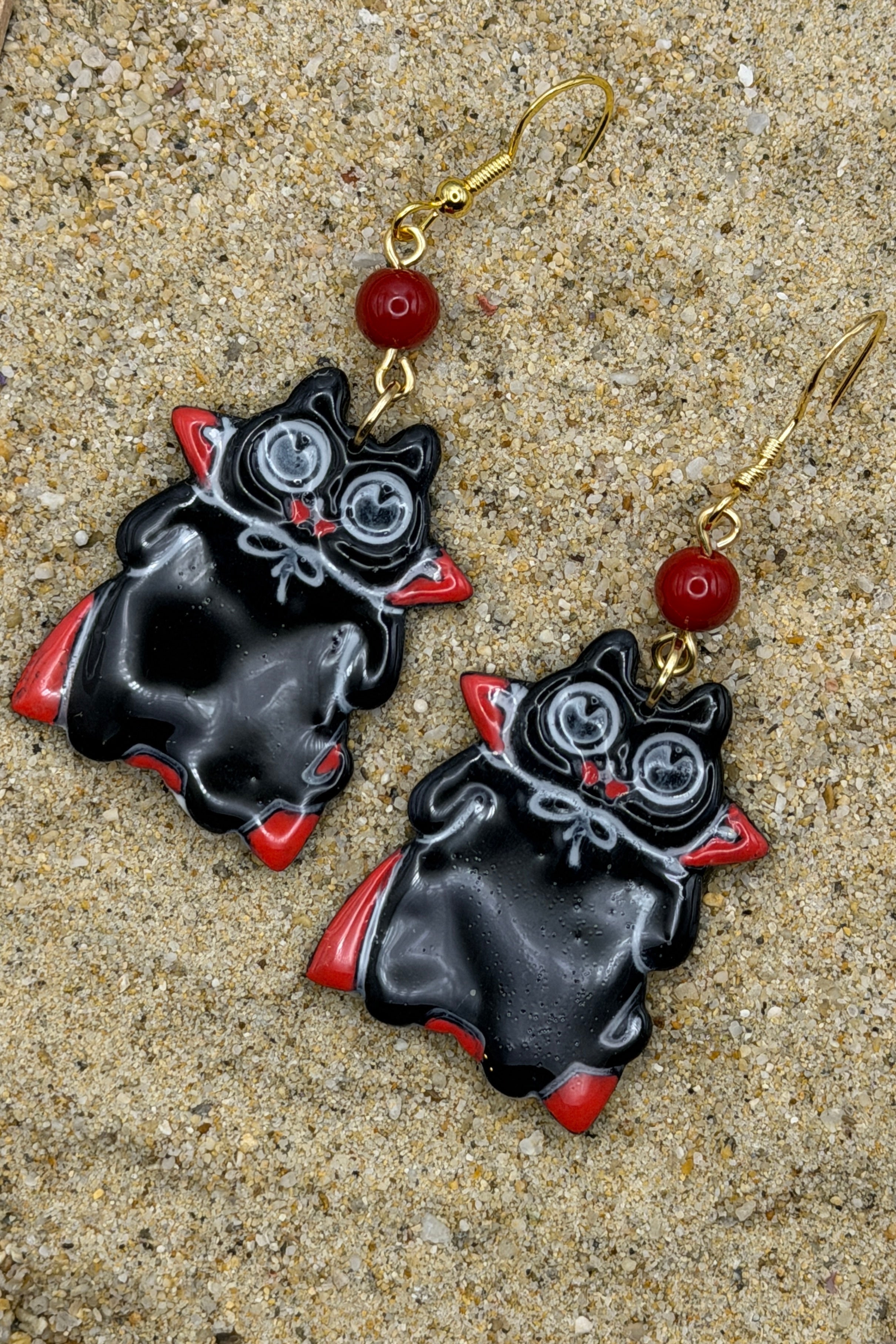 A pair of black vampire-shaped dangle earrings with red accents, silver eyes, and gold fishhook wires, displayed on a sandy surface.