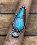 A sterling silver ring featuring a large turquoise stone and a scalloped sunburst accent, displayed on a wooden dowel over a rustic wood plank background.