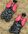 A pair of dangling earrings laid on sandy ground, featuring black skull-and-crossbones shapes with bright pink bows and small black heart details at the top.