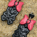 A pair of dangling earrings laid on sandy ground, featuring black skull-and-crossbones shapes with bright pink bows and small black heart details at the top.