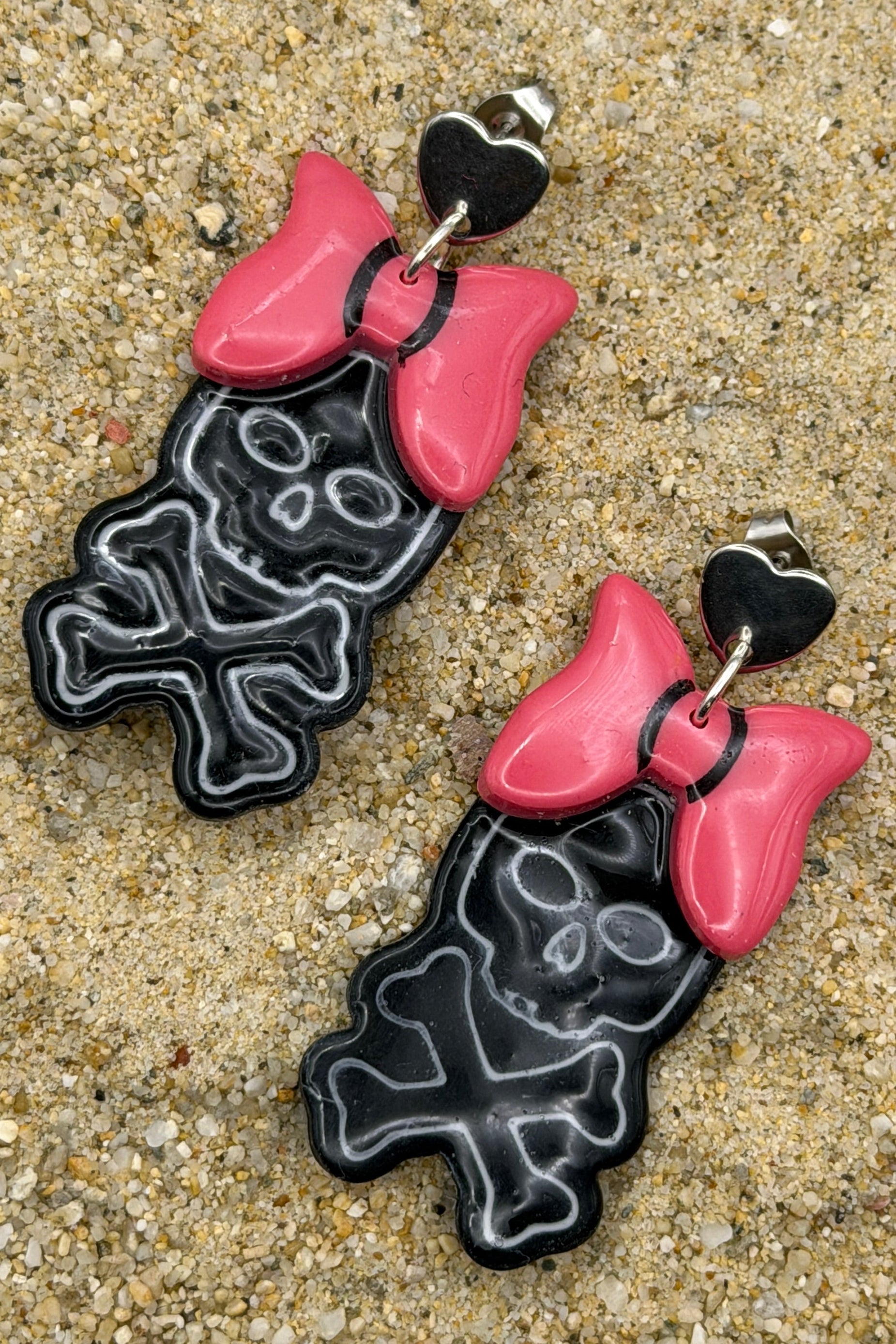 A pair of dangling earrings laid on sandy ground, featuring black skull-and-crossbones shapes with bright pink bows and small black heart details at the top.