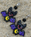 A pair of bat-shaped dangle earrings with purple wings, red eyes, and yellow crescent moons, displayed on a sandy surface.