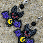 A pair of bat-shaped dangle earrings with purple wings, red eyes, and yellow crescent moons, displayed on a sandy surface.