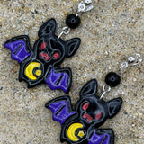 A pair of bat-shaped dangle earrings with purple wings, red eyes, and yellow crescent moons, displayed on a sandy surface.