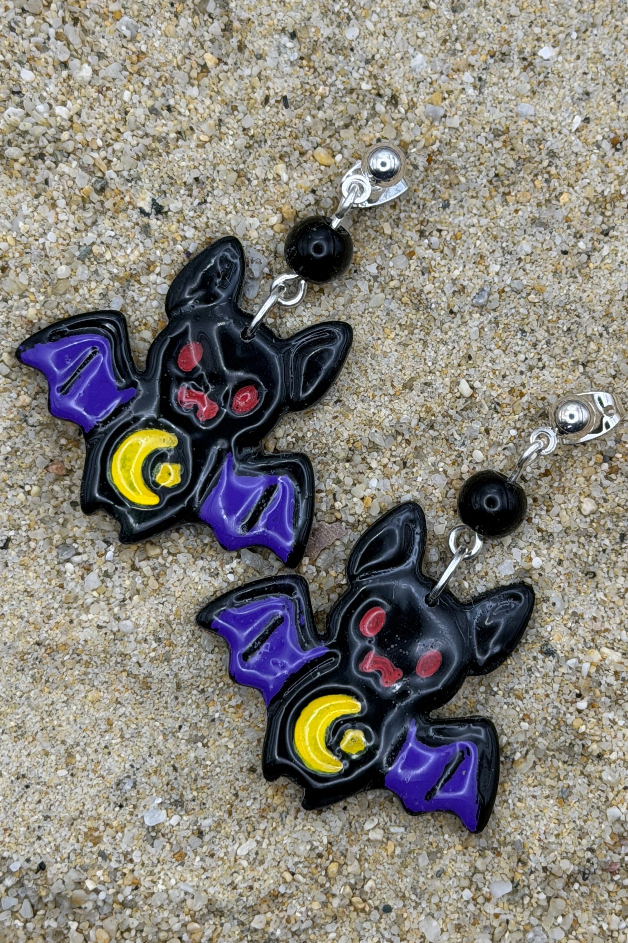 A pair of bat-shaped dangle earrings with purple wings, red eyes, and yellow crescent moons, displayed on a sandy surface.