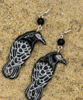 A pair of dangling raven earrings with black and white patterned bodies, small red eyes, and black beads, displayed on a sandy surface.
