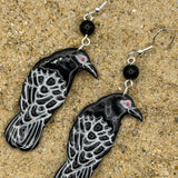 A pair of dangling raven earrings with black and white patterned bodies, small red eyes, and black beads, displayed on a sandy surface.
