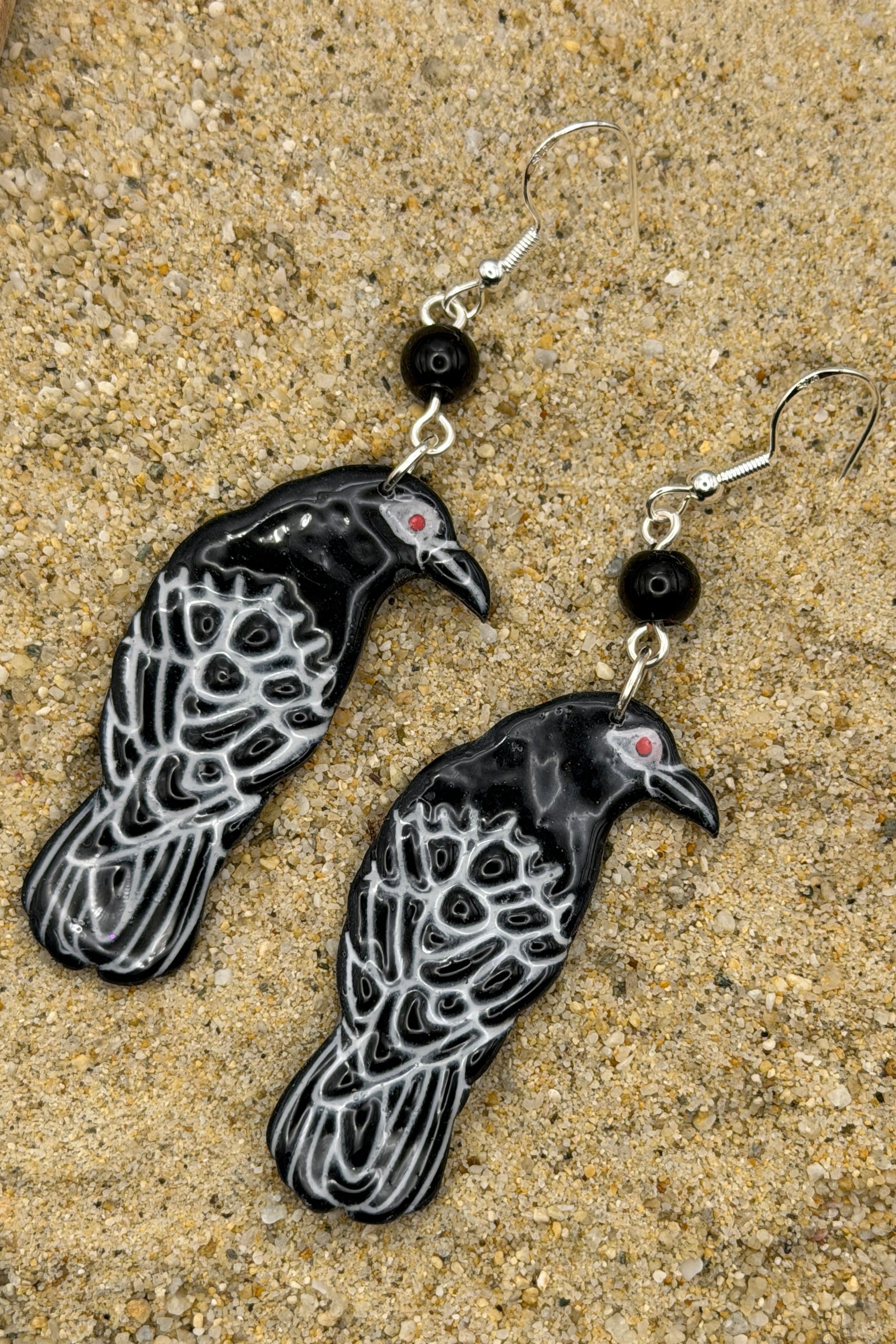A pair of dangling raven earrings with black and white patterned bodies, small red eyes, and black beads, displayed on a sandy surface.