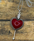 A silver heart pendant necklace with a red stone center and arrow detail, displayed on a rustic wooden surface.