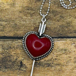 A silver heart pendant necklace with a red stone center and arrow detail, displayed on a rustic wooden surface.