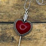 A silver heart pendant necklace with a red stone center and arrow detail, displayed on a rustic wooden surface.