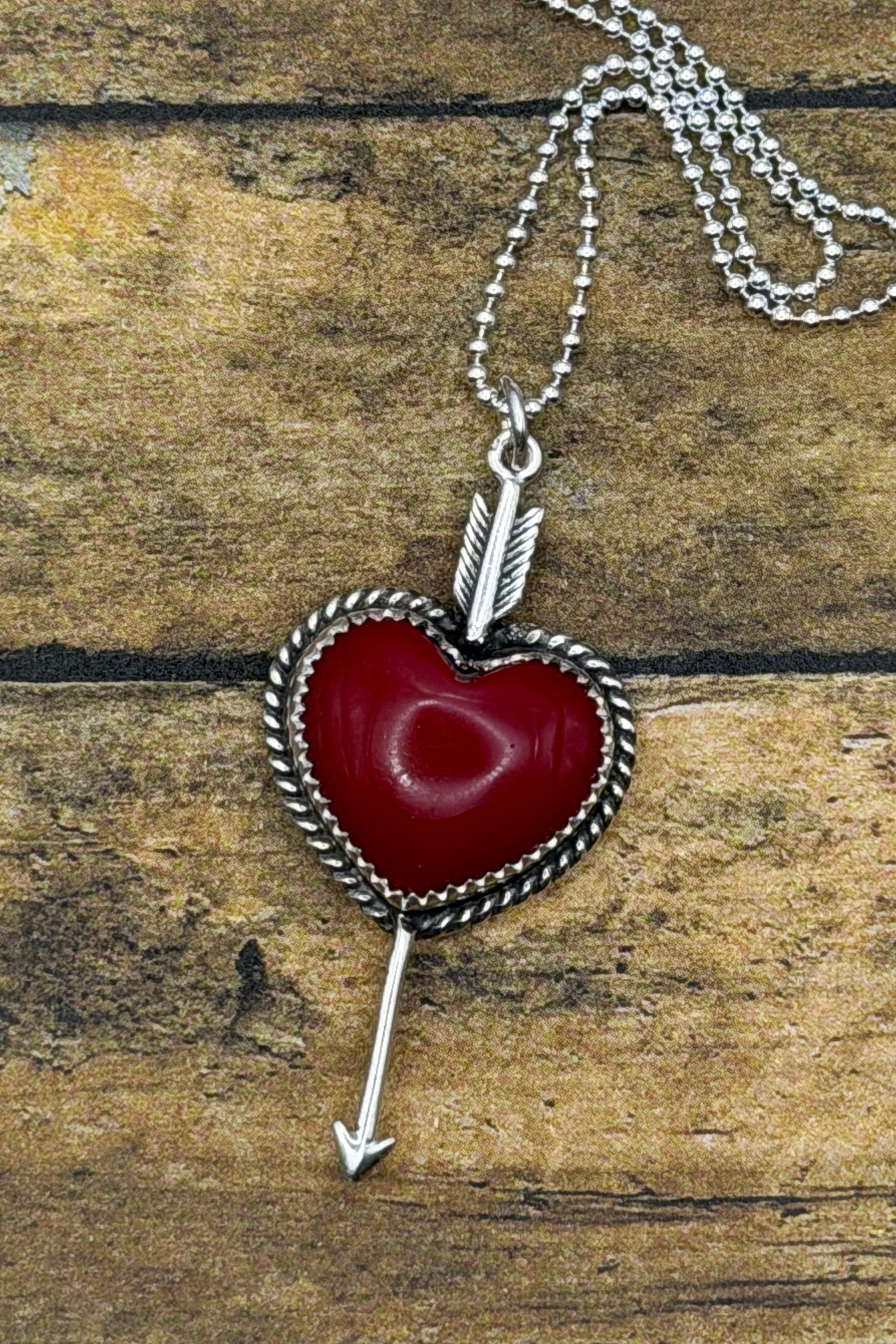 A silver heart pendant necklace with a red stone center and arrow detail, displayed on a rustic wooden surface.