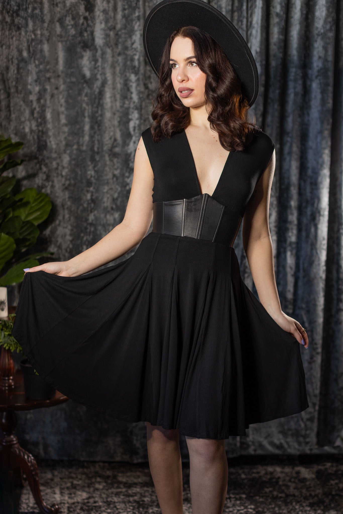 Woman wearing a black dress with a leather corset detail against a textured gray curtain background.