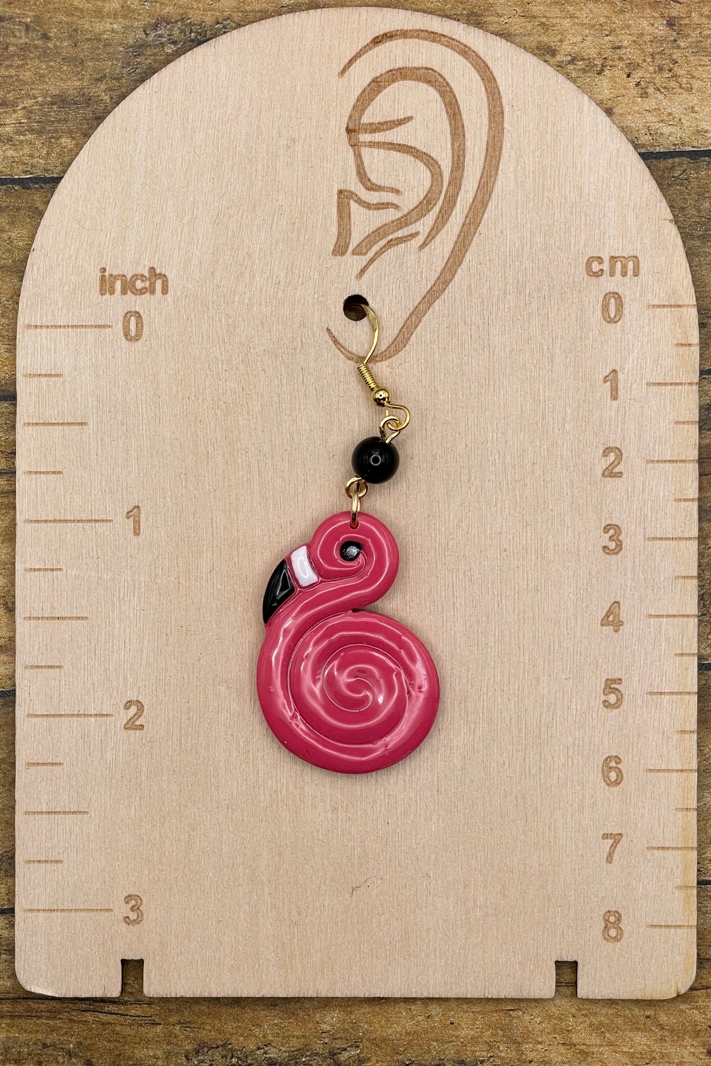 Pink spiral flamingo earring on a wooden measurement chart with inch and cm scales.