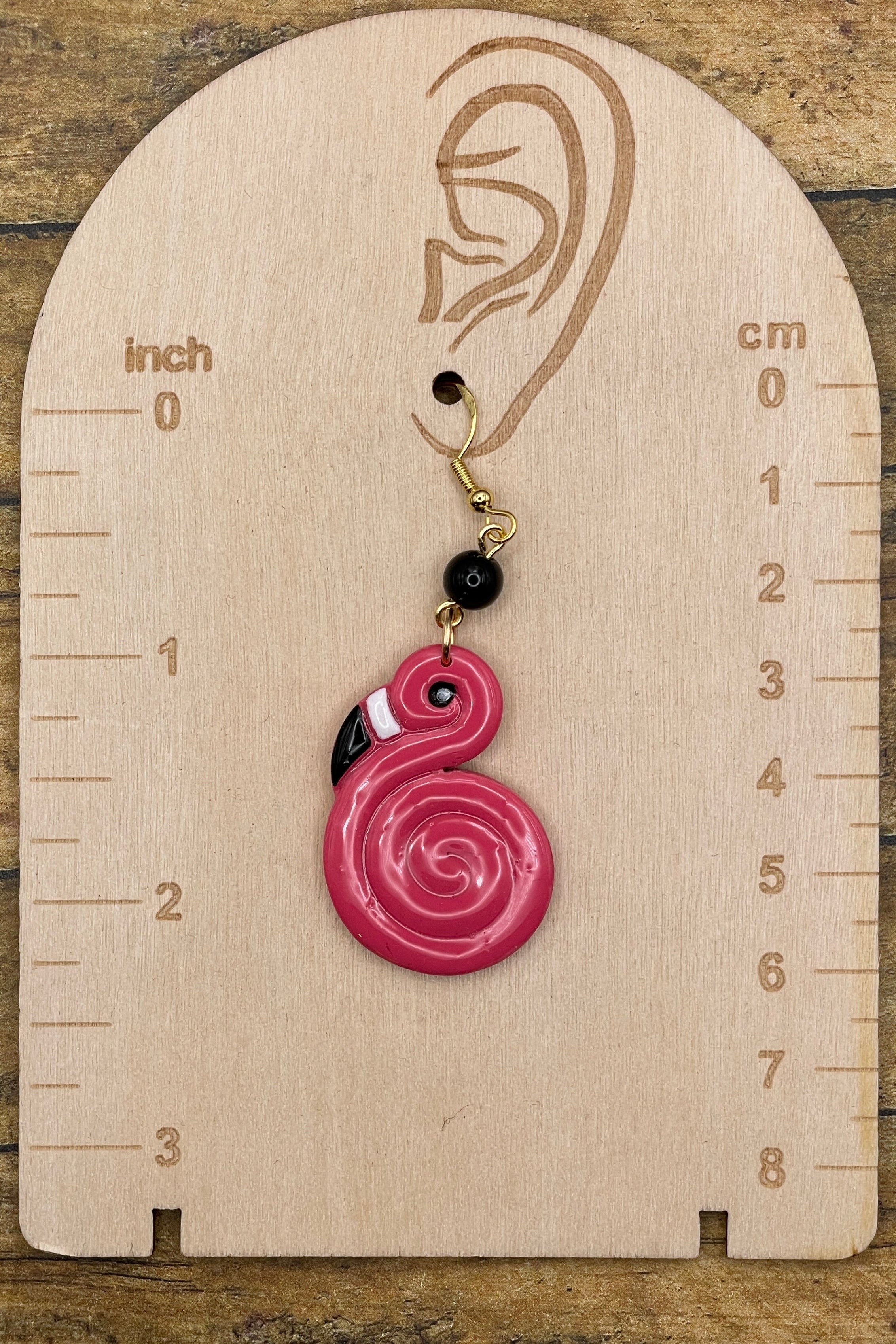 Pink spiral flamingo earring on a wooden measurement chart with inch and cm scales.